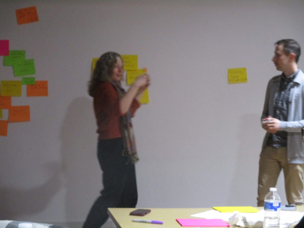 Helen (on the left) and Arran (on the right) stand in front of grouped together post-it notes, gesturing to and reading out suggestions for a conference North Star.