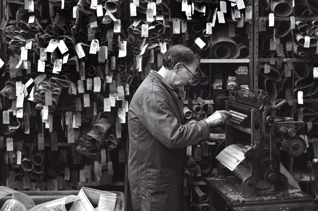 A man stands in front of machine programming it to punch cards. Behind they are stacks of rolled, punched paper, all labelled. In the foreground is basket of waste paper. 