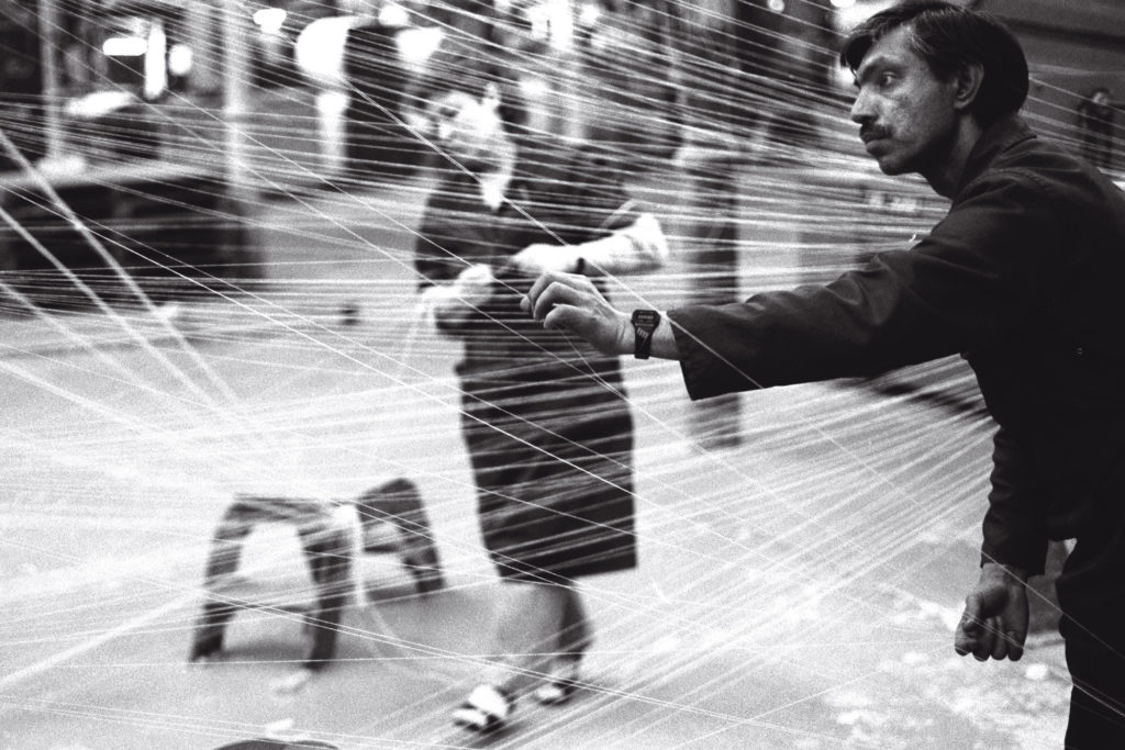 A photo of two people, a man and a women, either side of warp for weaving. The strands of material spread out, criss-crossing in front of the photographer.