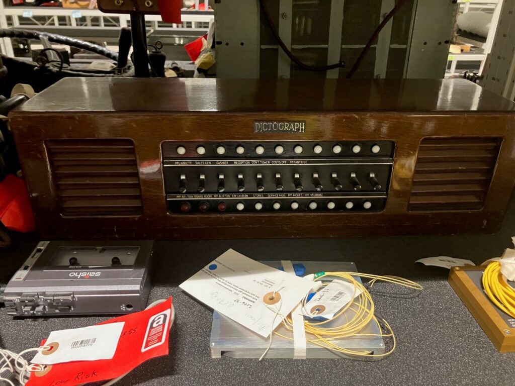 A desk on which an object on it, this is an early intercom with flick switches on the front. Wires and bits of paper are on the table around it. 